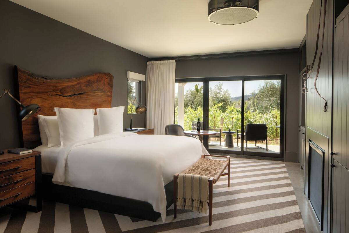 Ein modernes Hotelzimmer mit einem großen Bett, einem Holz-Kopfteil, einem Schreibtisch mit Stuhl, einem kleinen Tisch mit Stuhl und Blick ins Grüne durch große Fenster.