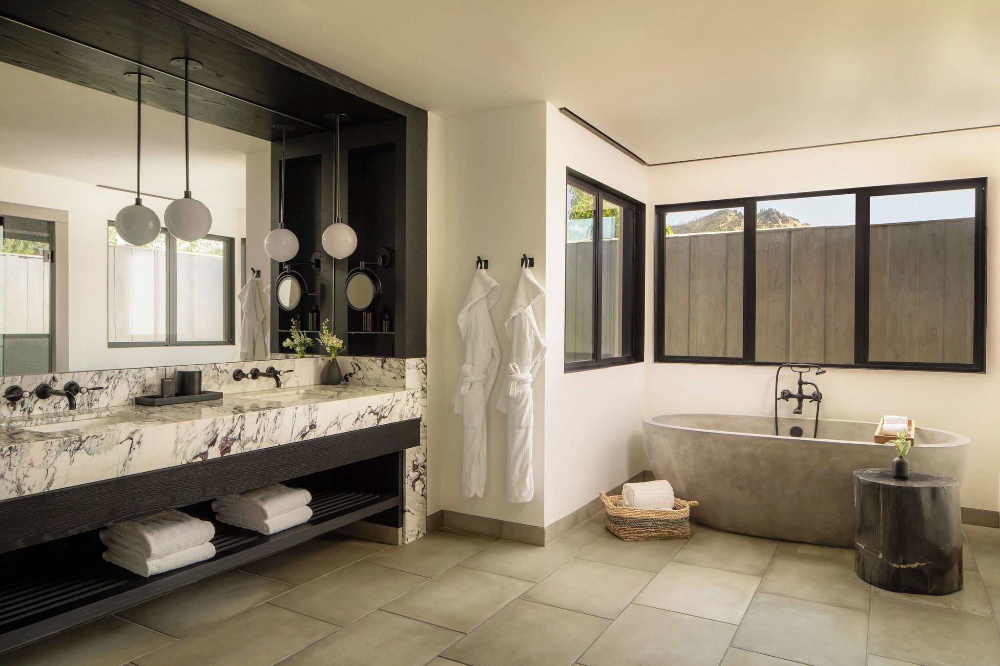 A modern bathroom with a large mirror, a marble vanity, a freestanding bathtub, and a window with a view of the outdoors.