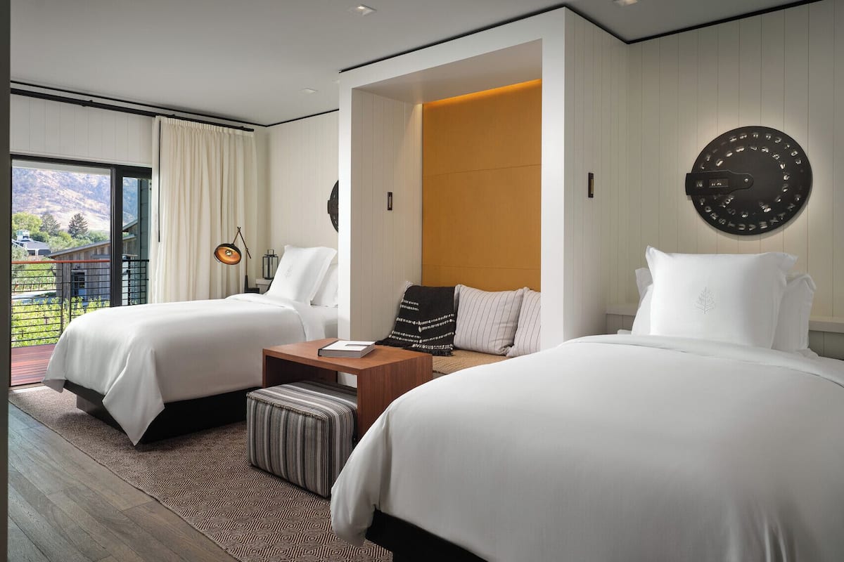 Ein modernes Hotelzimmer mit zwei Betten, einem großen Fenster mit Vorhängen, einer Uhr an der Wand und einem kleinen Tisch mit einer Lampe.