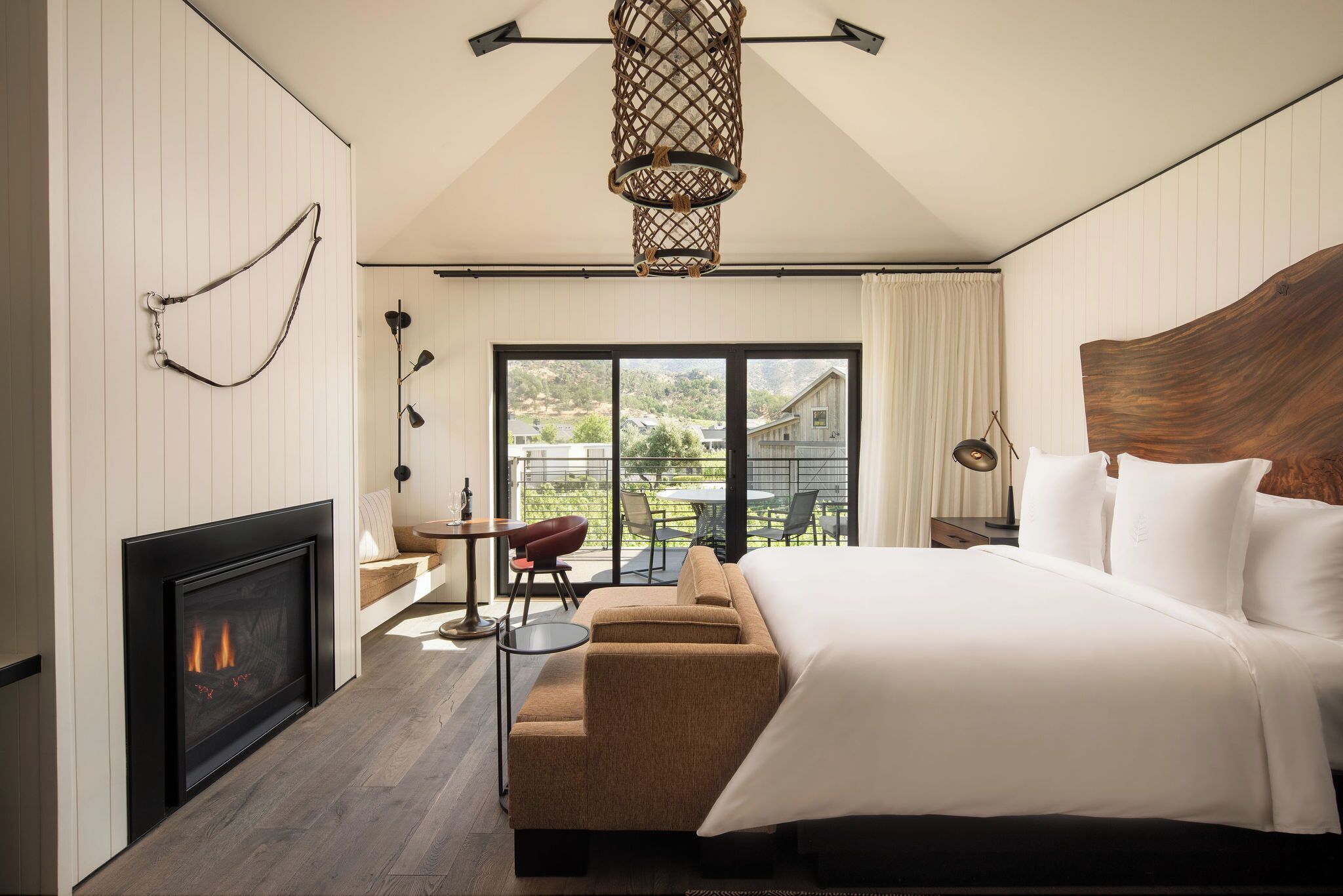 A modern bedroom with a fireplace, a large bed, a sofa, and a balcony with a view of the countryside.