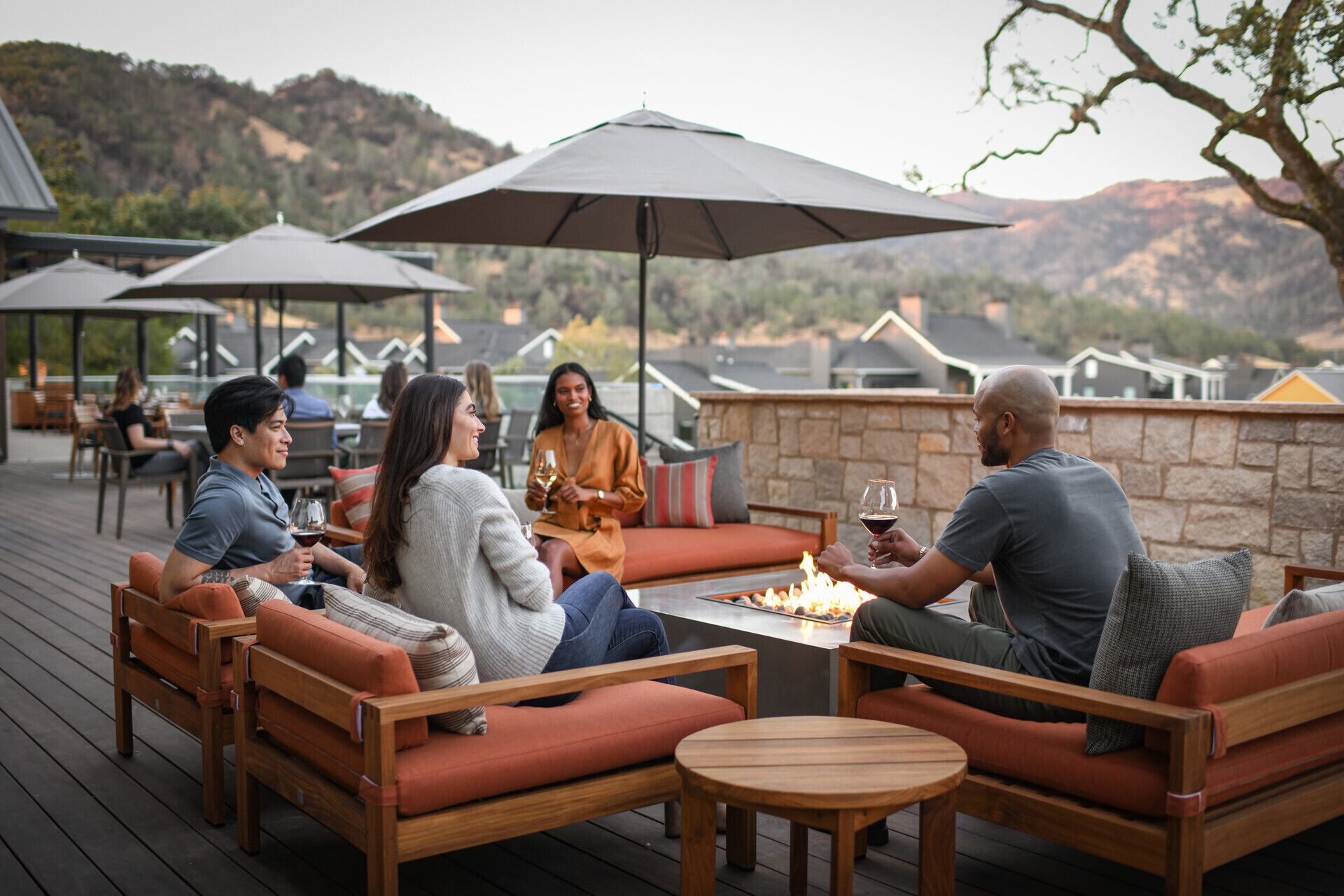 A group of people seated at a wooden table with a fire pit, surrounded by outdoor furniture and umbrellas, overlooking a mountainous landscape.
