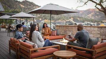 A group of people seated at a wooden table with a fire pit, surrounded by outdoor furniture and umbrellas, overlooking a mountainous landscape.