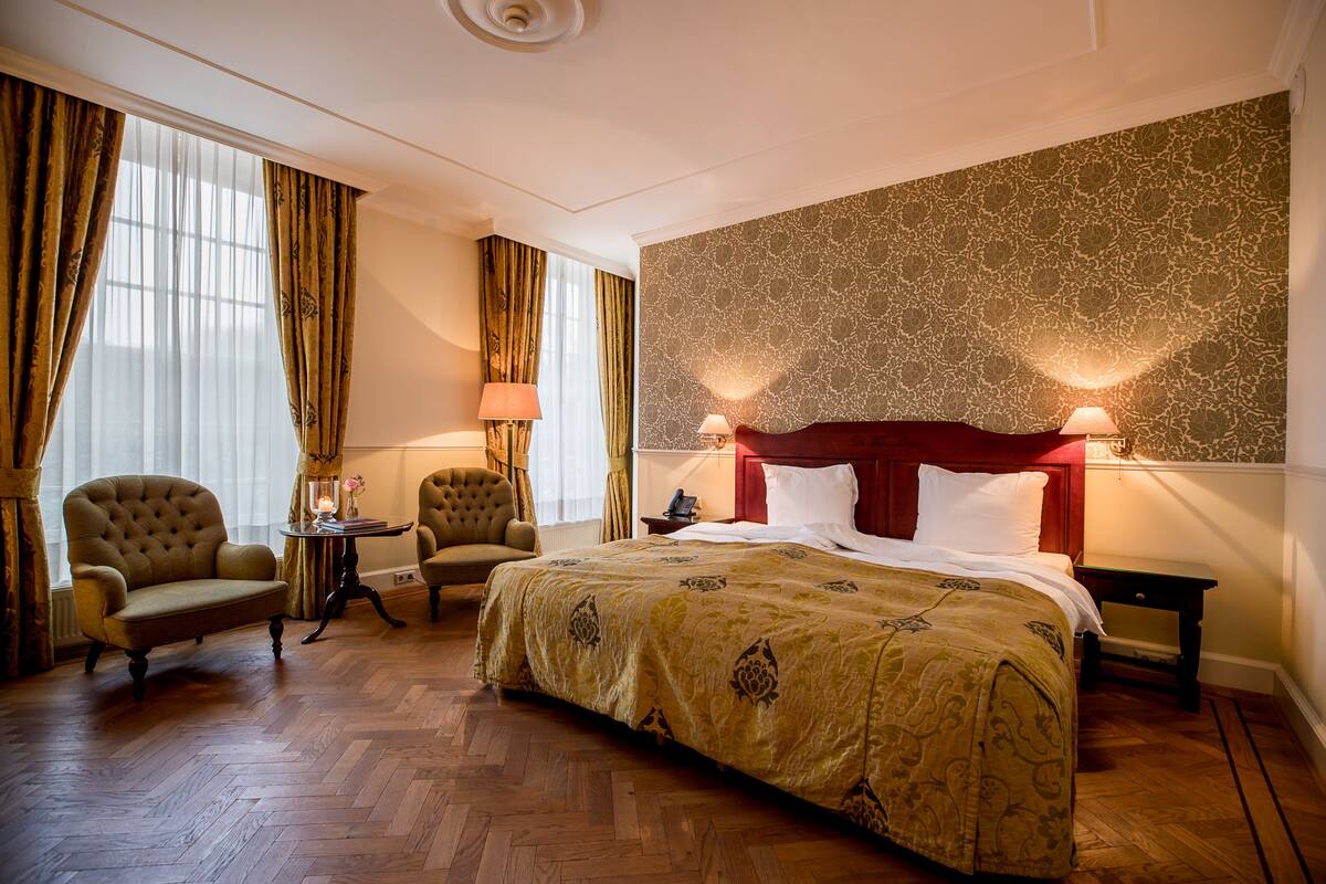 Une chambre d’hôtel avec un grand lit, deux fauteuils, une petite table avec une lampe et un mur décoré d’un motif.