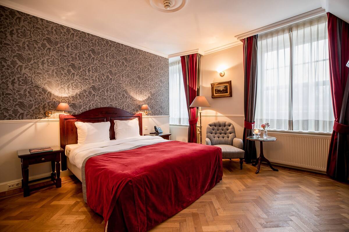 Une chambre d’hôtel avec un grand lit, une chaise, une petite table et une fenêtre avec des rideaux.