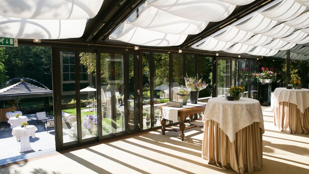 A covered outdoor area with tables and chairs, large windows, and a view of a garden. - Kasteel de Wittenburg (Wassenaar)
