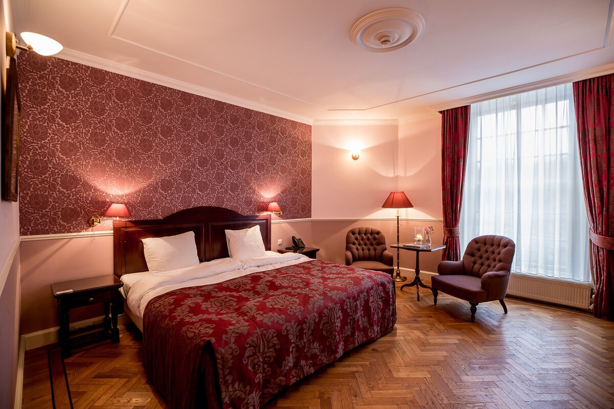 Een hotelkamer met een groot bed, een zitstoel, een tafeltje en een raam met gordijnen.
