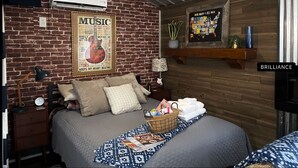 Een knus ingerichte kamer met een bakstenen muur, een poster van een gitaar, een bed met een bedekt dekbed, een bedlamp en een klein plankje met een plantje.