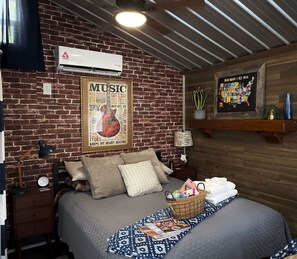 Een knus ingerichte kamer met een bakstenen muur, een poster van een gitaar, een bed met een bedekt dekbed, een bedlamp en een klein plankje met een plantje.