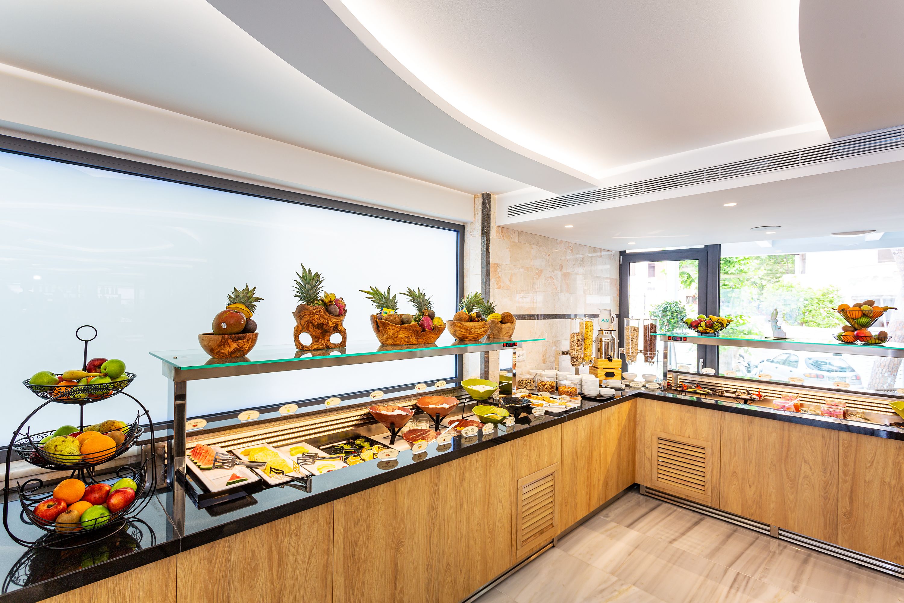 En buffet med forskelligt frugt og mad, en glasmontre og udsigt til det fri gennem store vinduer.