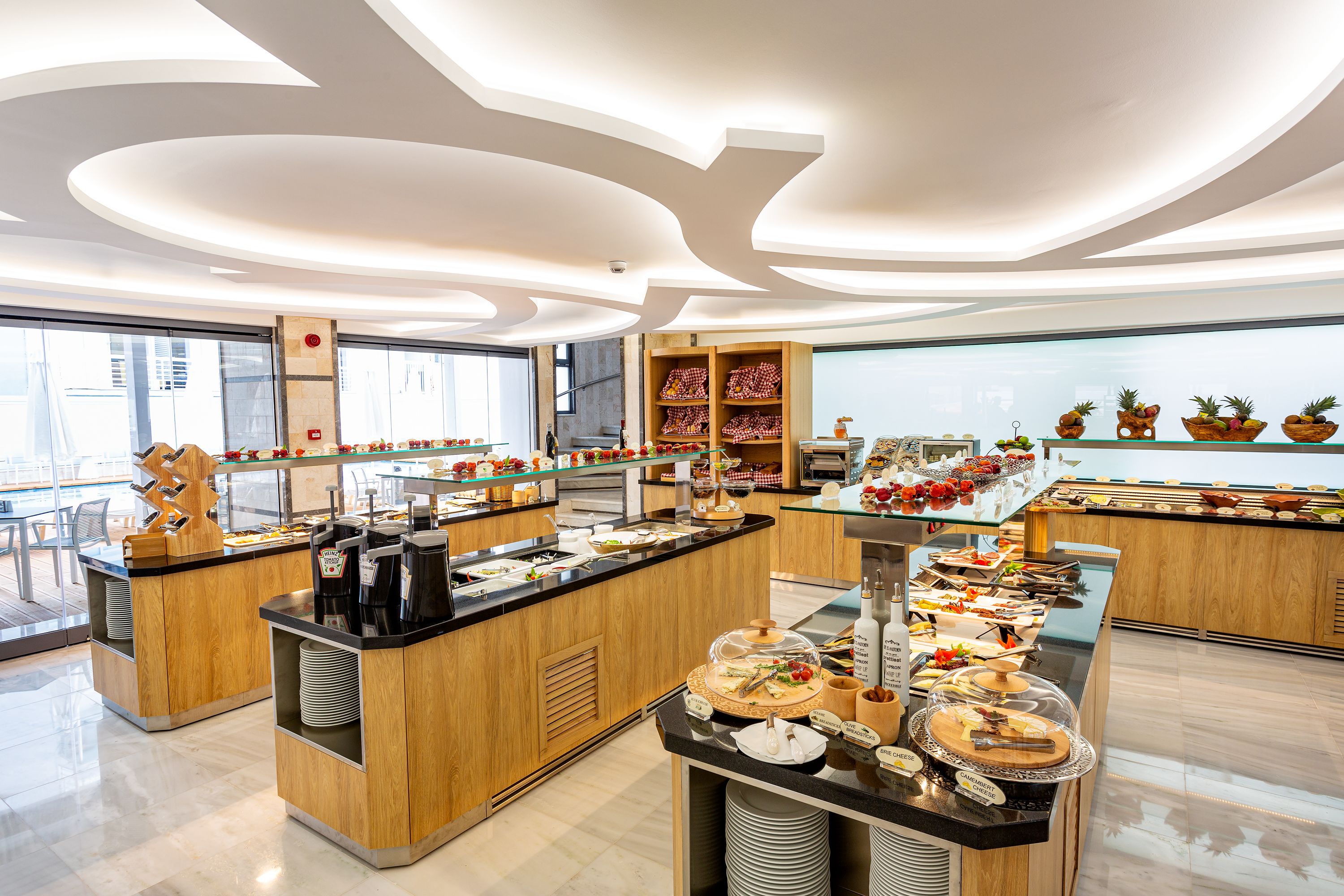 Un moderno bufé de desayuno con múltiples estaciones de servicio, una gran variedad de alimentos y un diseño espacioso con ventanas amplias.