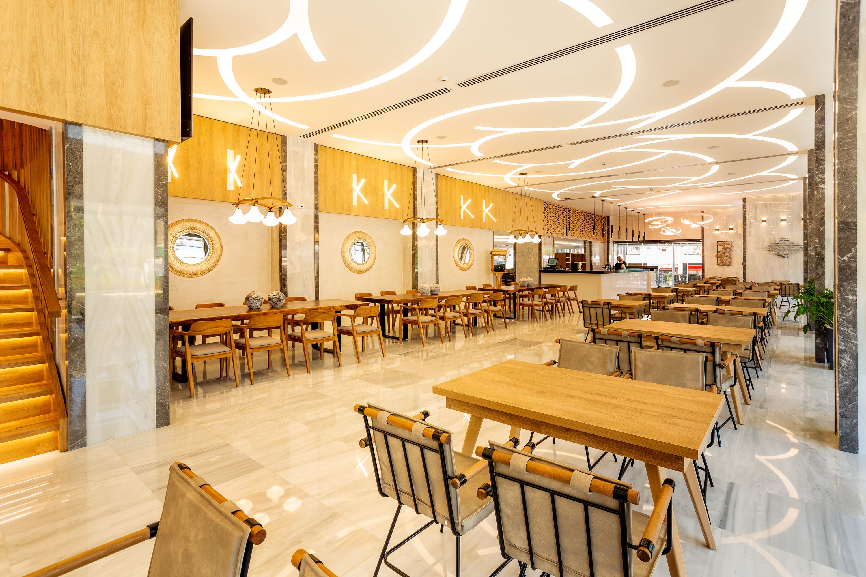 A spacious dining area with multiple wooden tables and chairs, a modern ceiling design, and a visible counter area in the background.