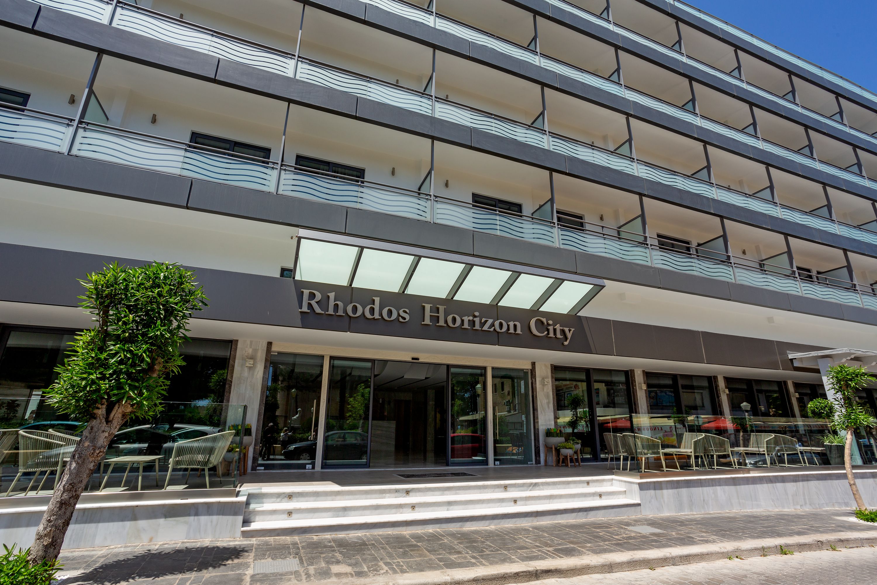 A modern building with a sign reading "Rhodos Horizon City." It features multiple balconies with glass railings and a glass entrance.