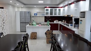Uma cozinha moderna com armários brancos, eletrodomésticos de aço inoxidável e uma ilha central. Há cadeiras ao redor de uma mesa de jantar e caixas no chão.