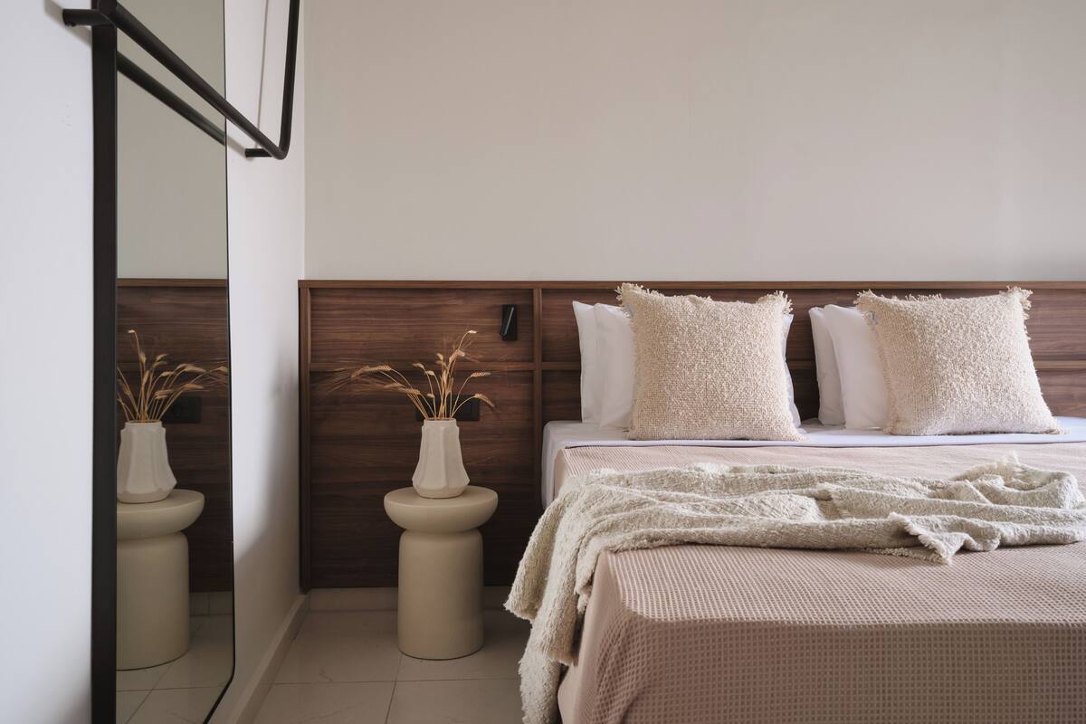 Un dormitorio con cabecera de madera, una cama con ropa de cama blanca, dos mesitas de noche y un espejo en la pared.