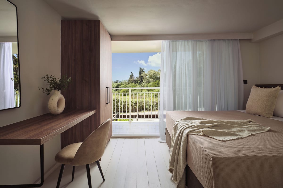 Un dormitorio con cama, escritorio, silla, planta en maceta y balcón con vista a follaje.