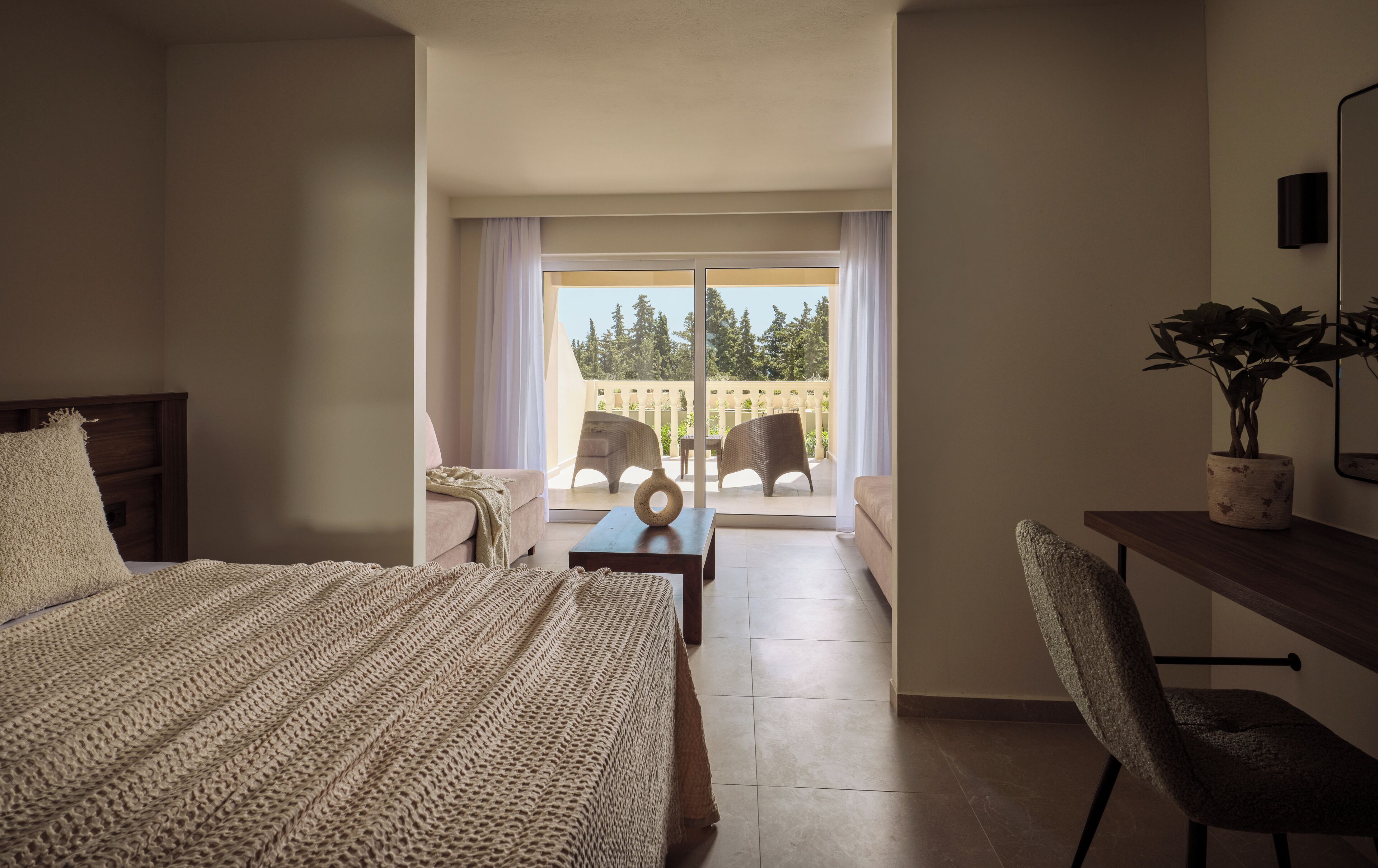 A hotel room with a bed, a desk, a chair, a small table, and a balcony with outdoor furniture.