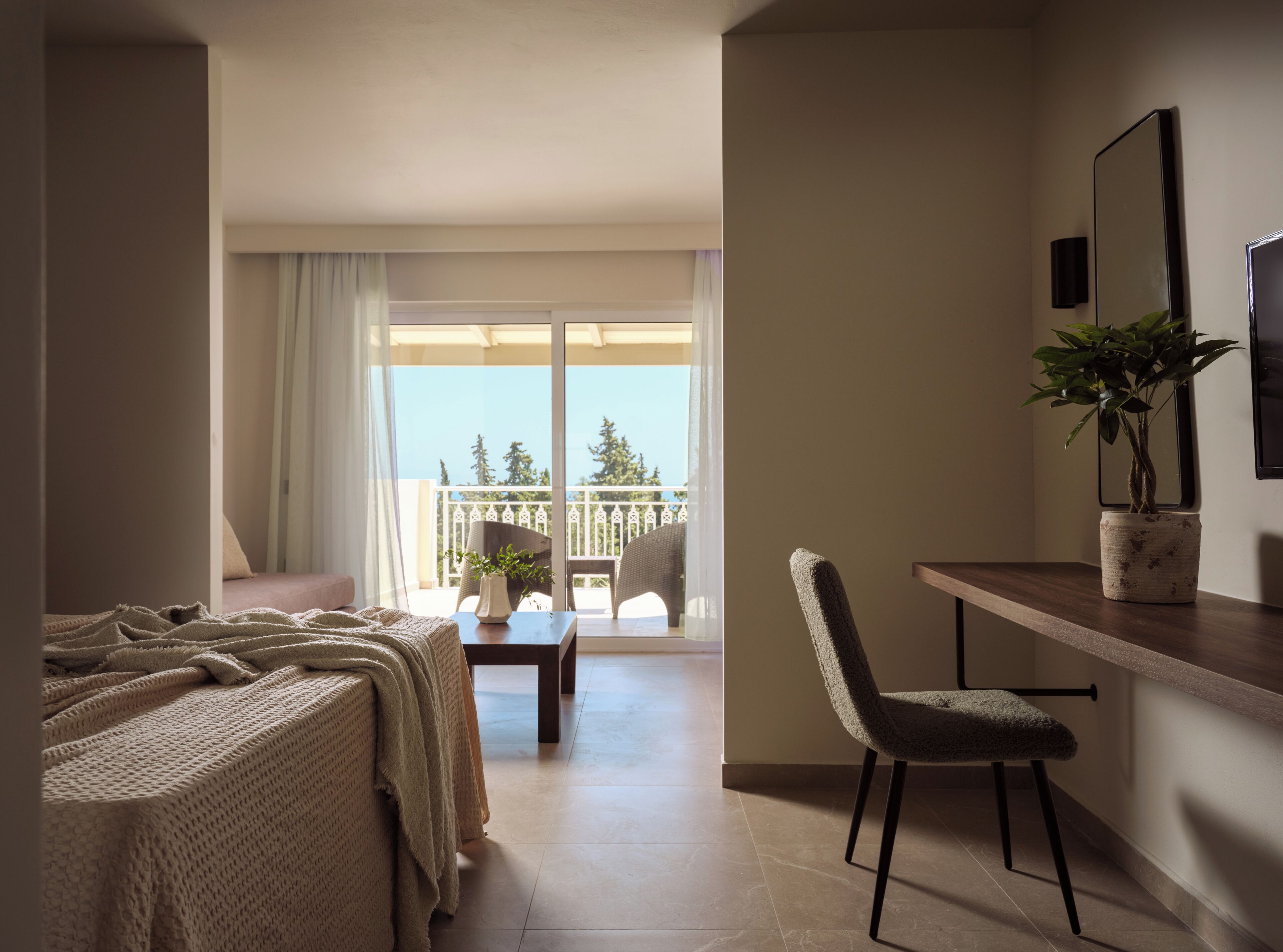 A hotel room with a bed, a desk, a chair, a potted plant, and a balcony with a view of trees.