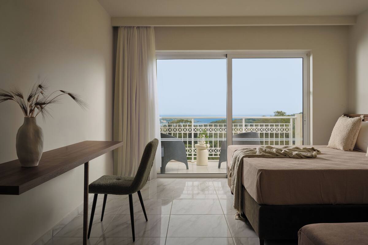 Habitación de hotel con cama, escritorio, una silla, una planta en maceta y un balcón con vistas.