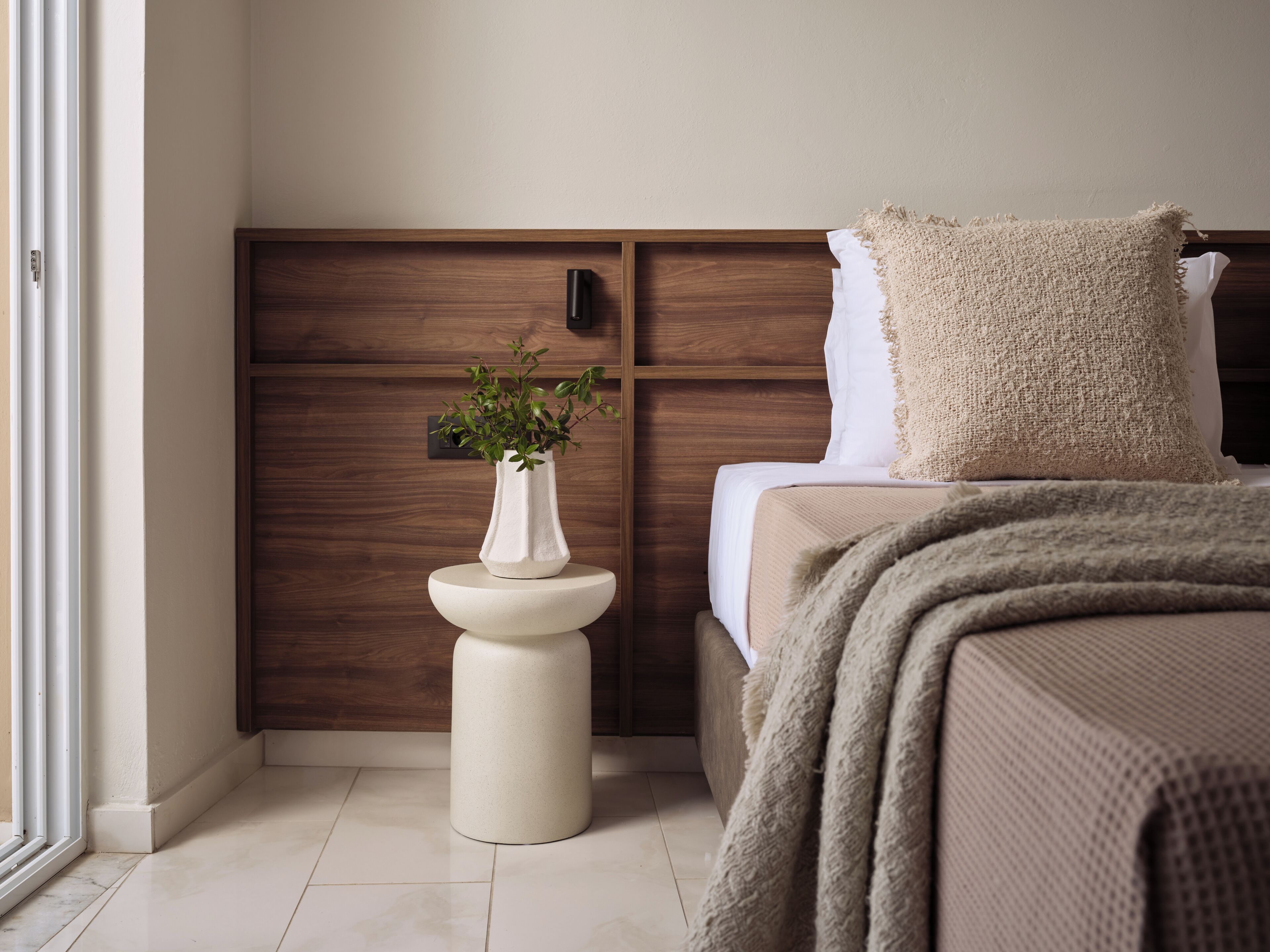 A bedroom with a wooden headboard, a bedside table with a white vase, a bed with a beige blanket, and a textured pillow.