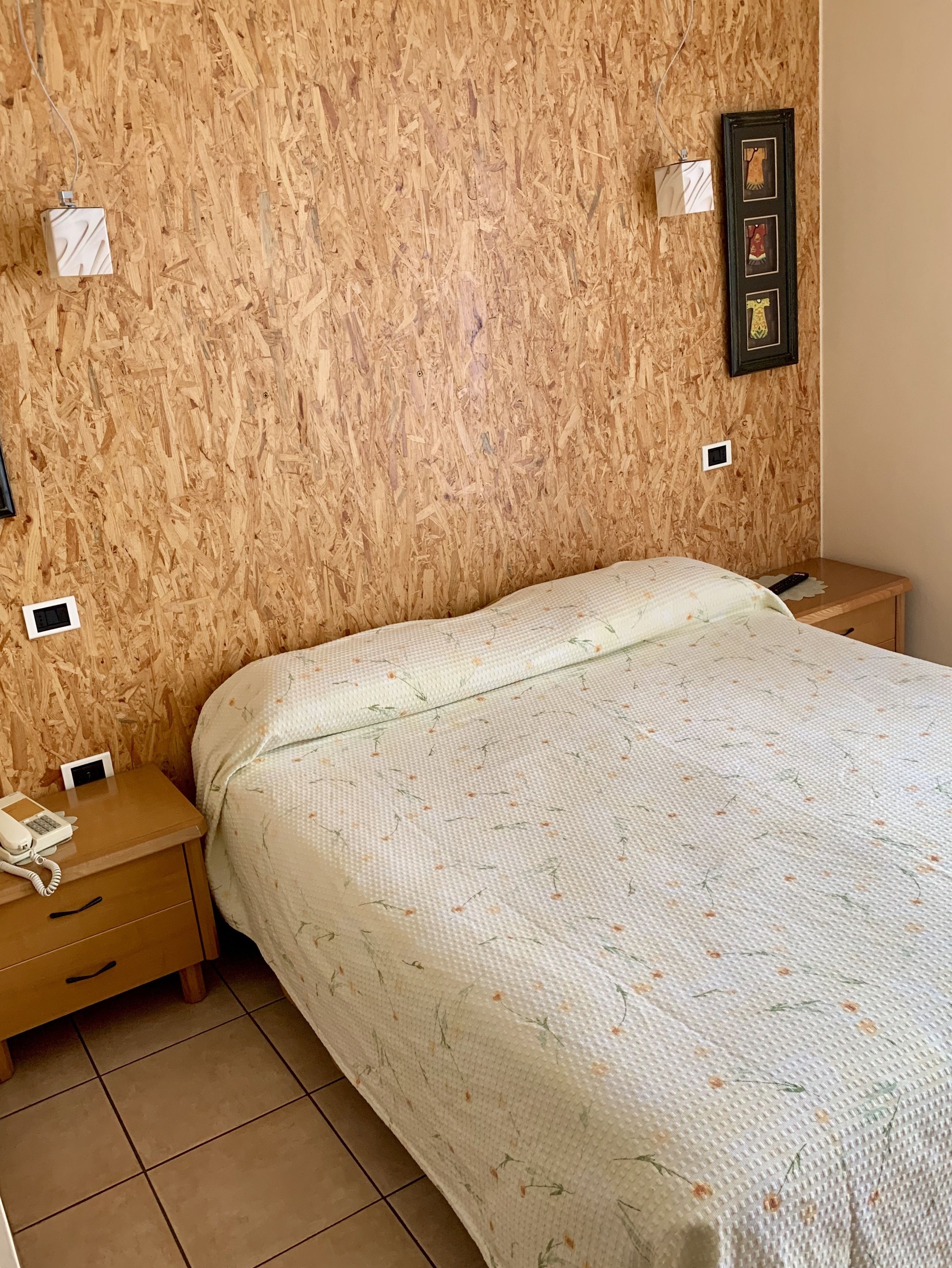 Un dormitorio con cabecera de madera, cama con colcha estampada, dos mesitas de noche con lámparas y un cuadro enmarcado en la pared.