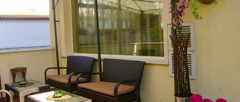 Un patio cubierto con muebles de mimbre, una mesa de vidrio y una maceta con flores rosadas.