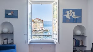 A room with a large window overlooking a waterfront with buildings and boats. There are two wall-mounted shelves with books and decorative items. - JOIVY La Finestra Sulla Piazzetta a Portofino (Portofino)