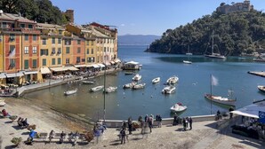 A waterfront promenade with buildings, boats, and a hill with a castle-like structure. - JOIVY La Finestra Sulla Piazzetta a Portofino (Portofino)