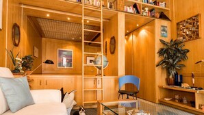 Ein gemütliches Wohnzimmer mit einer Couch, einem Glastisch, einem blauen Sessel und einem Hund. Holzregale sind mit Büchern und dekorativen Objekten gefüllt, es gibt einen Globus und eine große Pflanze.