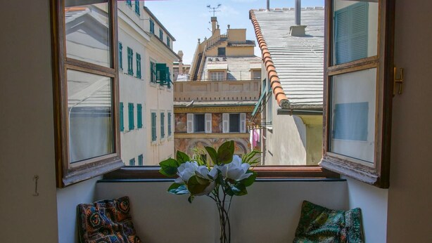 Janela com vista para uma rua estreita, um radiador embaixo e duas almofadas coloridas em um banco.