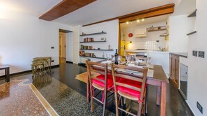Uma sala de jantar com uma mesa e cadeiras de madeira, uma estante de livros e uma cozinha visível através de uma porta aberta.