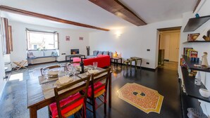 Ampla sala de estar com uma mesa de jantar posta para quatro pessoas, um sofá vermelho e uma lareira.