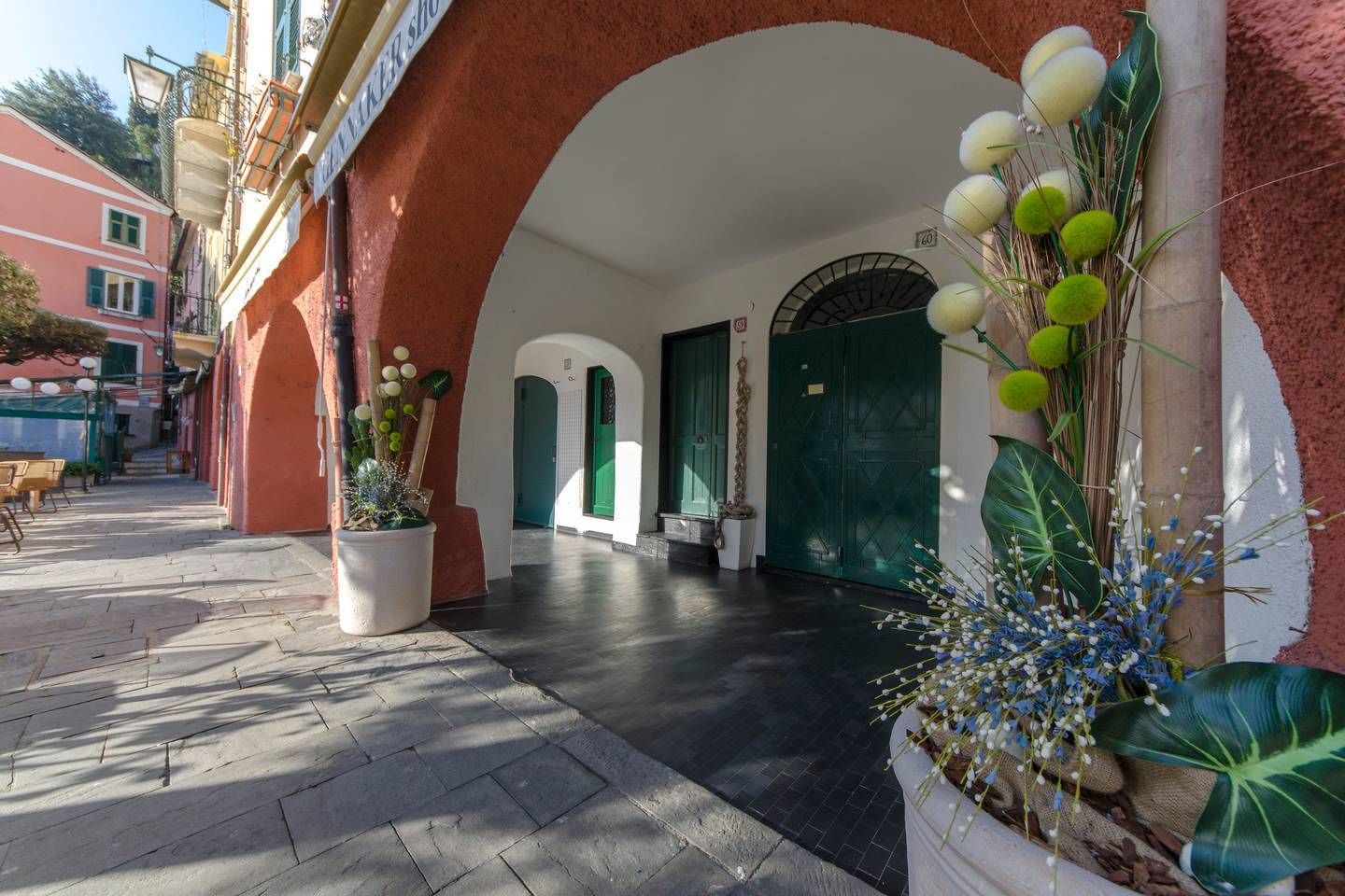 A hotel entrance with a red facade, white arches, and green doors. There are potted plants with white and green foliage.