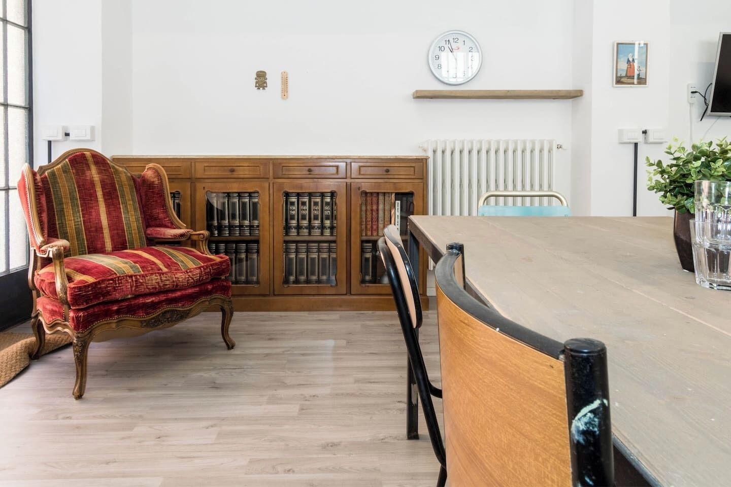A dining area with a wooden table and chairs, a red upholstered chair, a radiator, a clock, and a shelf with books.