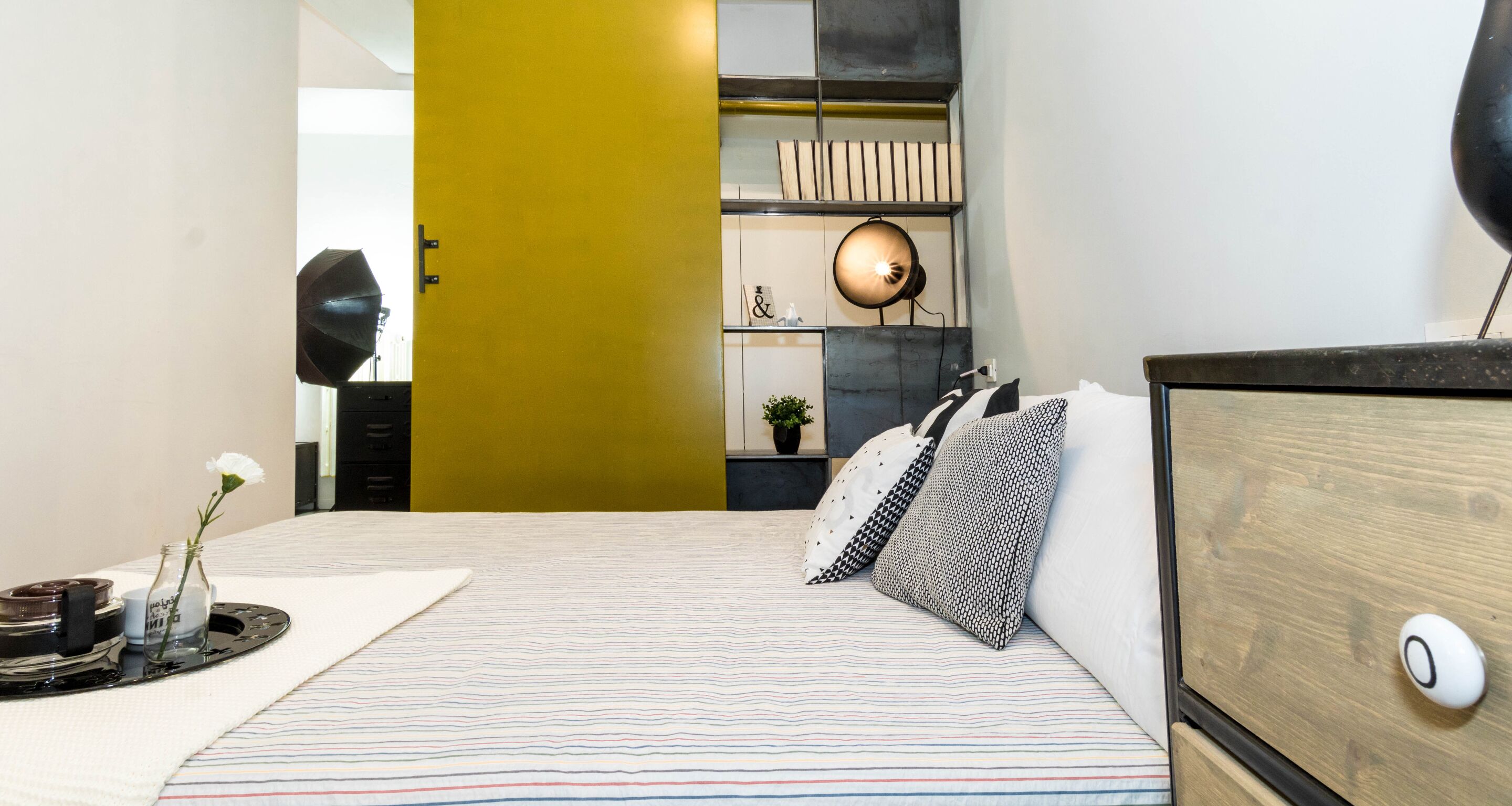 A bedroom with a bed, a bedside table, a lamp, and a shelf with decorative items.