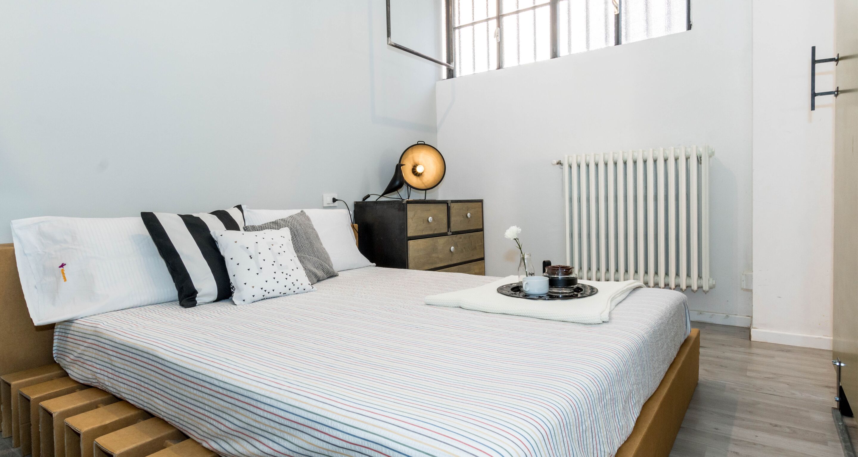 Un lit bien fait, avec une literie à rayures, une table de chevet avec une lampe, un radiateur et une fenêtre avec des barreaux.