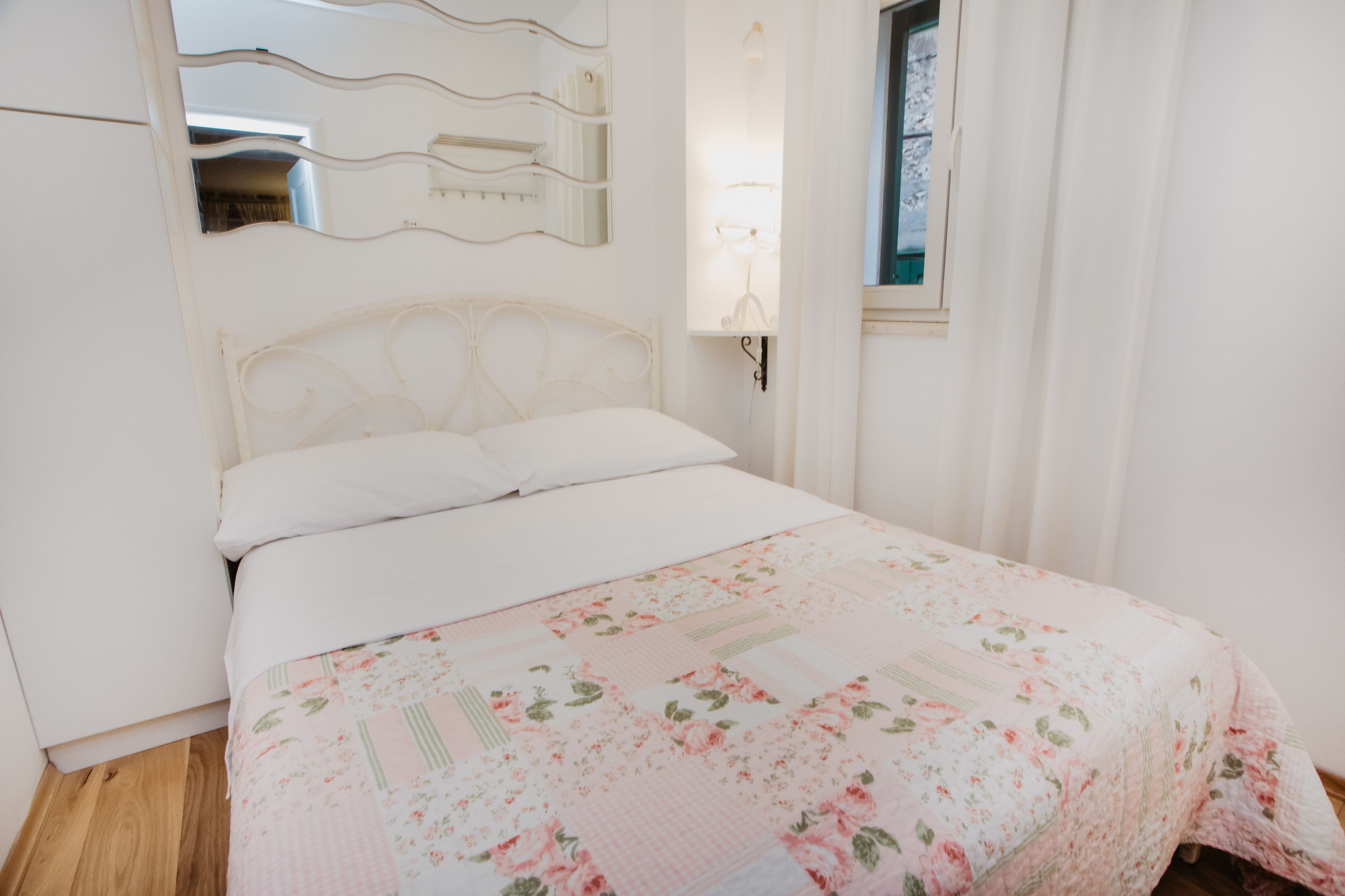 A bedroom with a floral bedspread, a white headboard, a bedside table with a lamp, and a window with curtains.