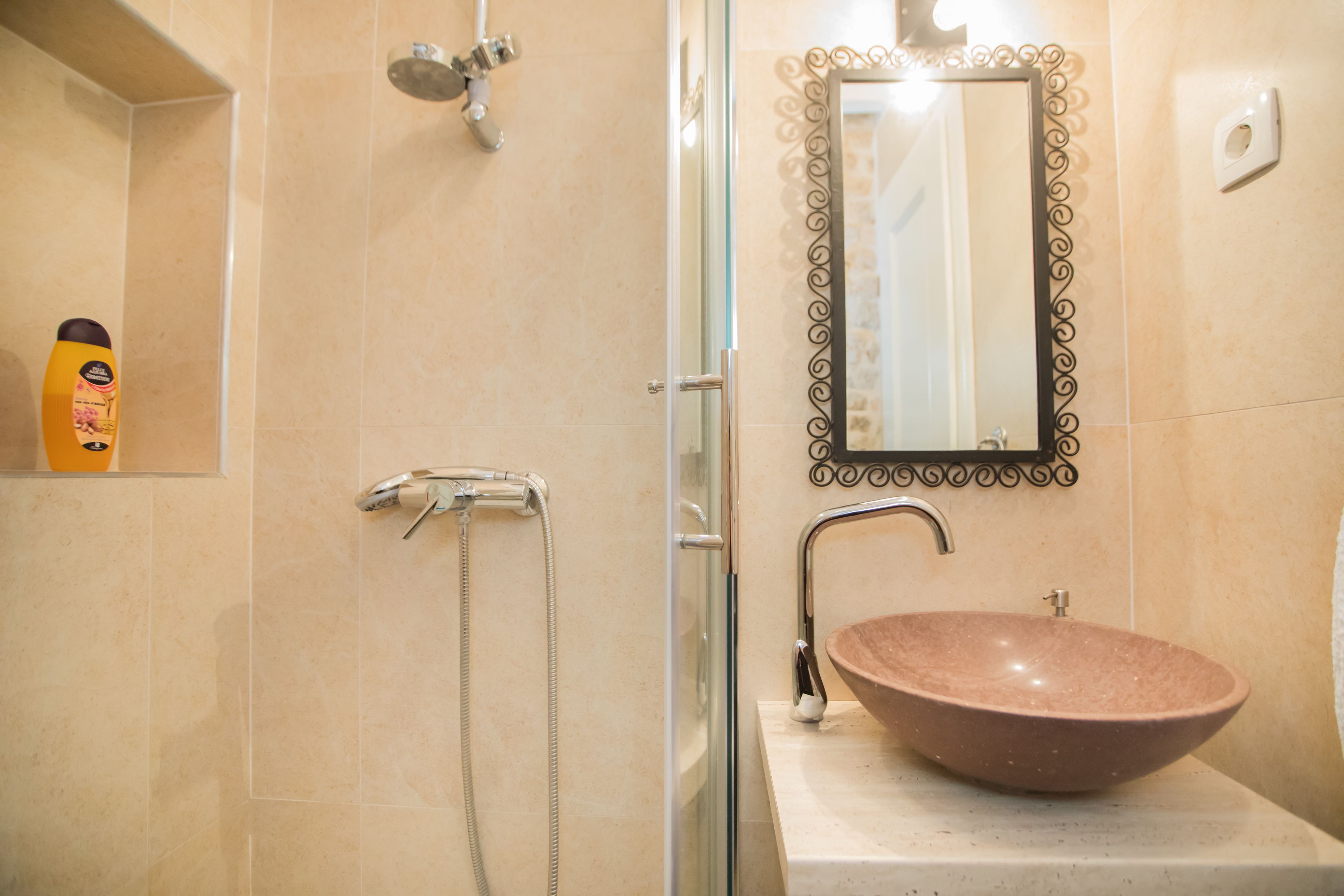A bathroom with a glass-enclosed shower, a beige tiled wall, a brown ceramic sink, and a decorative mirror with a black frame.
