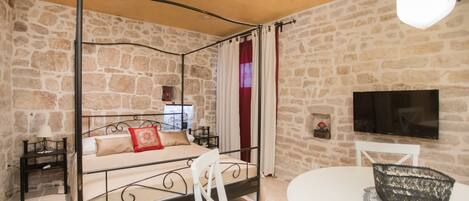 Una habitación con una cama con dosel, una silla blanca, una mesa redonda y un televisor montado en la pared.