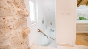 Un escalier, avec un mur de pierre d’un côté et un mur blanc de l’autre, mène à une chambre à coucher où se trouvent un lit et un oreiller vert.