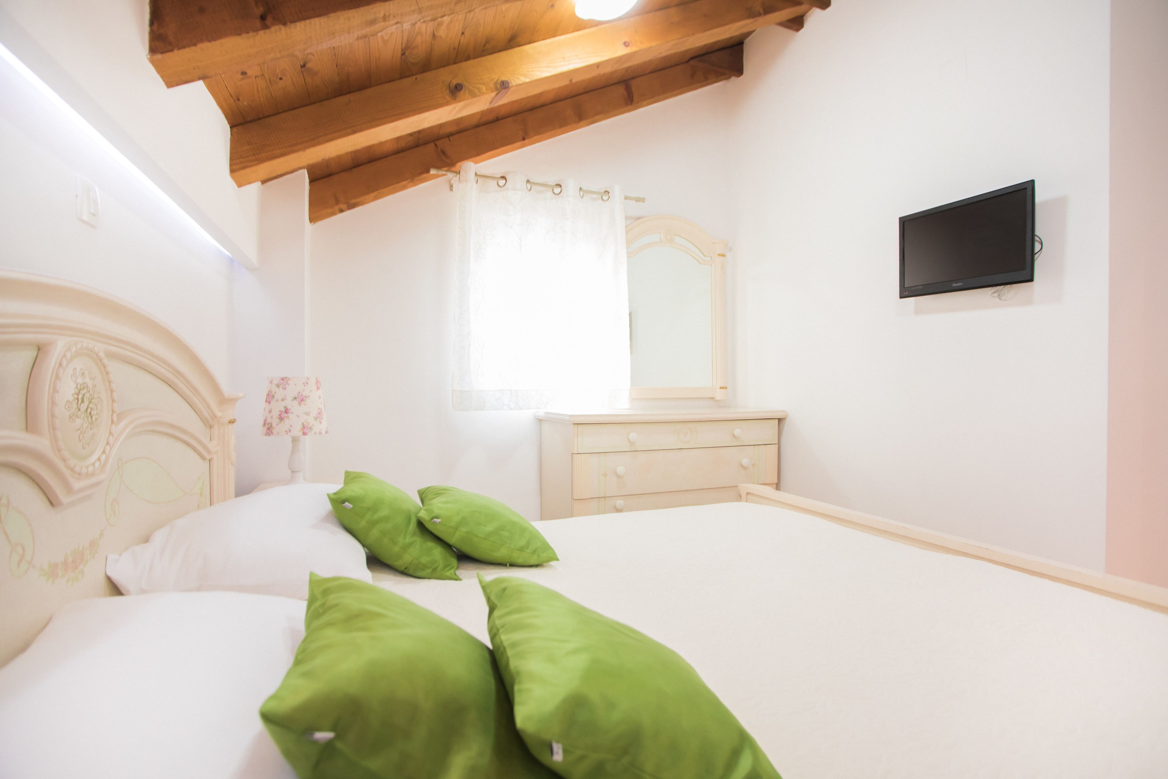 Un dormitorio con techo de madera, una cama con almohadas blancas y verdes, un aparador de madera, un espejo y un televisor de pantalla plana en la pared.