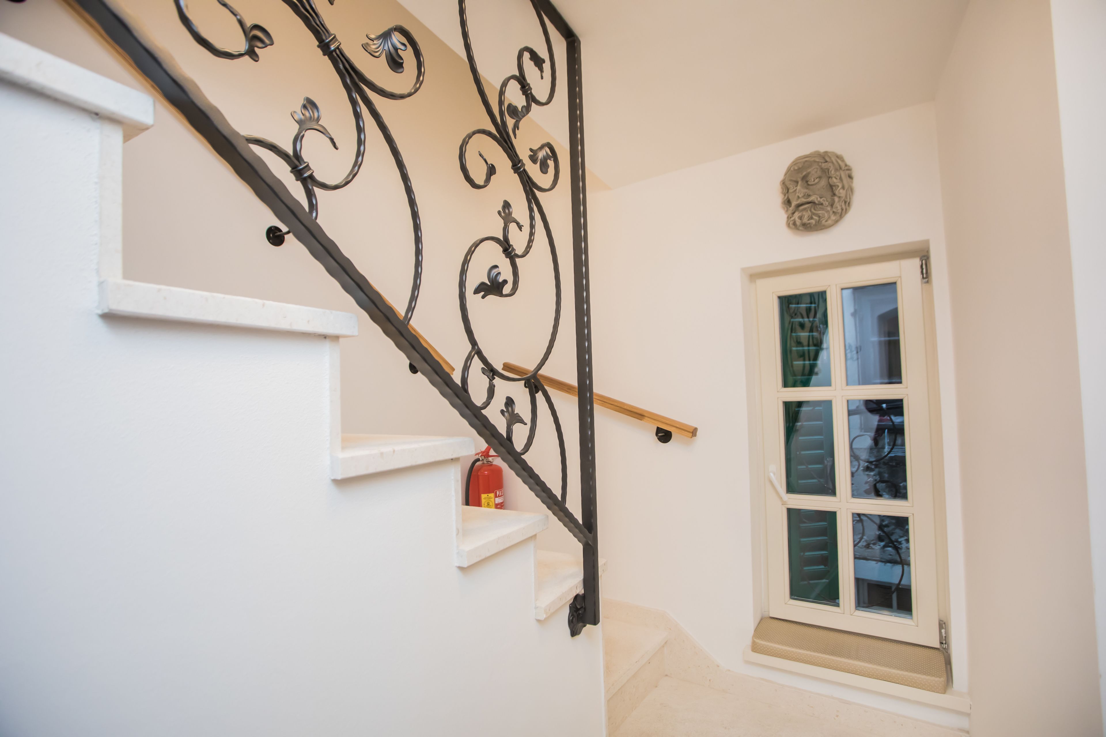 A staircase with a decorative iron railing, a white wall, a window with blinds, and a fire extinguisher on the wall.
