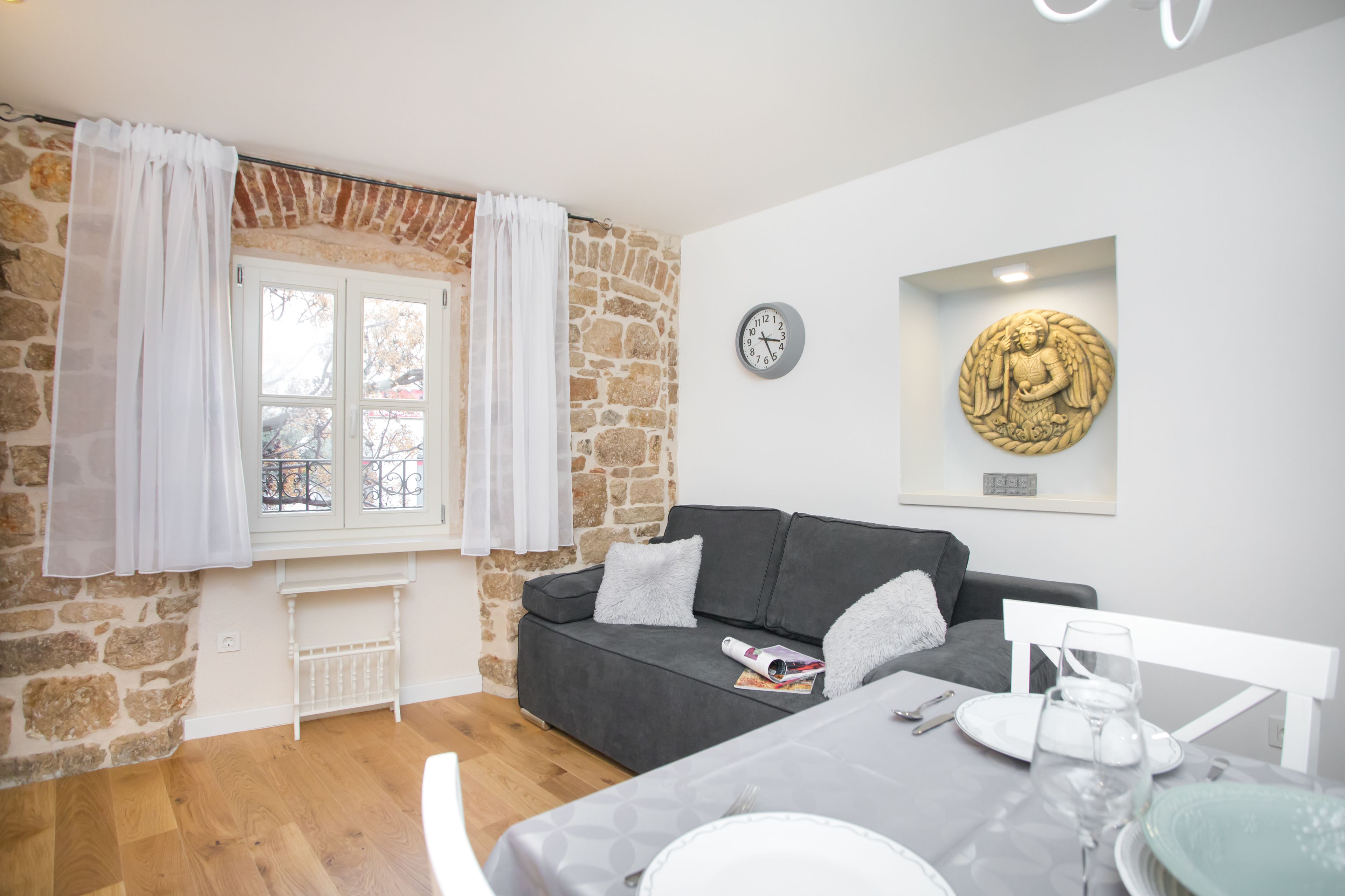 A cozy room with a stone wall, a grey sofa, a dining table set with plates and glasses, and a clock on the wall.
