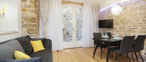 A living room with a grey sofa, yellow cushions, a dining table with chairs, a stone wall, and a TV mounted on the wall.