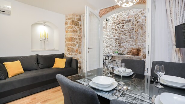 A modern living room with a black sofa, a dining table set for two, and a stone wall. - Residence Teatro - Near Cathedral of St James (Sibenik)