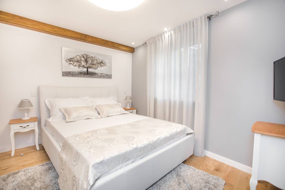 A bedroom with a large bed, white walls, a wooden ceiling beam, a painting of a tree, a TV on the wall, and a window with curtains.