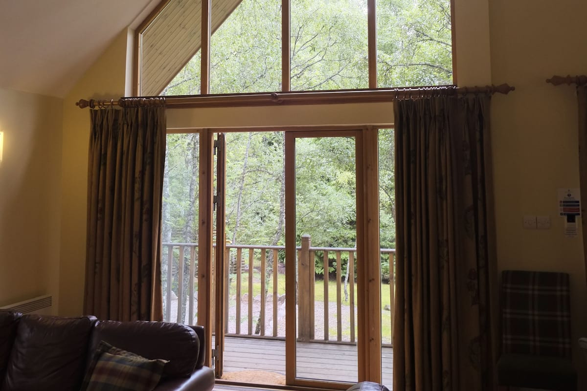 A spacious living room with a high ceiling, large windows, and a view of a wooden deck.