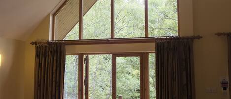 A spacious living room with a high ceiling, large windows, and a view of a wooden deck.