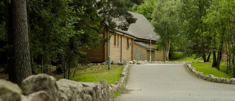 En hytte med træbeklædning og en asfalteret indkørsel, omgivet af træer og en stenvæg.
