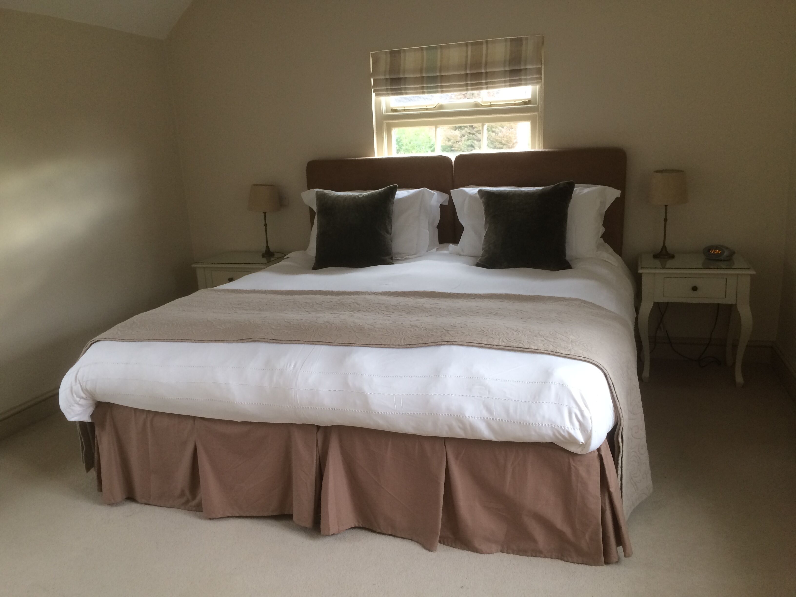 A neatly made bed with a brown skirt, two bedside tables with lamps, and a window with blinds.
