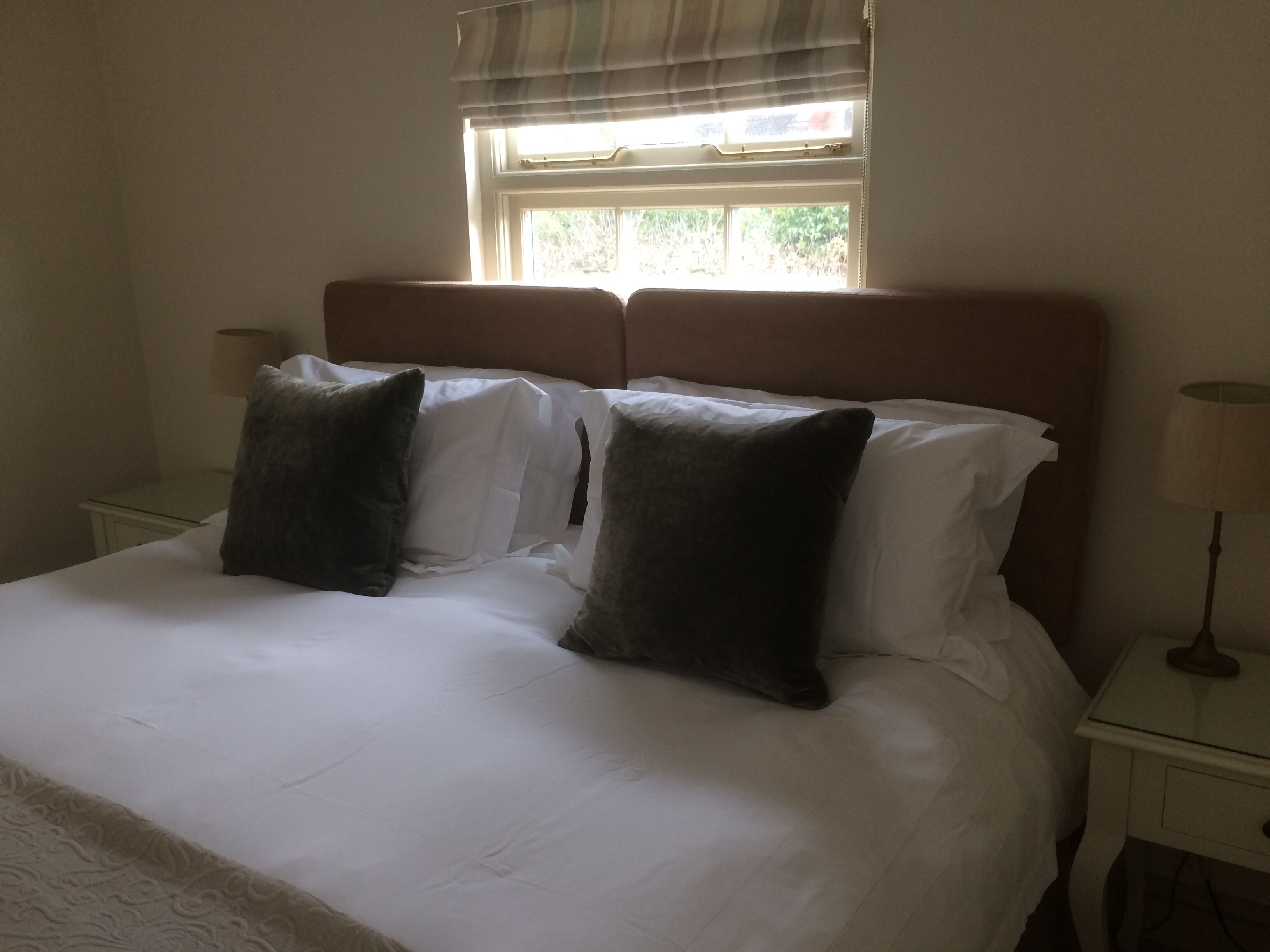 A neatly made bed with white linens and two dark pillows, a bedside table with a lamp, and a window with blinds.